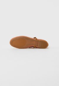 Suela de un zapato plano de ante rosa con un fondo de goma marrón texturizada y una pequeña hebilla de tira en el tobillo visible sobre un fondo blanco.
