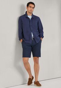 Námornícky modrá bunda na gombíky s vreckom na hrudi cez pruhovanú košeľu. V kombinácii s námorníckymi šortkami a hnedými semišovými slip-on topánkami. Hladká textúra látky.