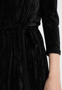 Vestido de terciopelo negro con textura plisada, mangas largas y una cintura atada, que enfatiza un acabado suave y lujoso.