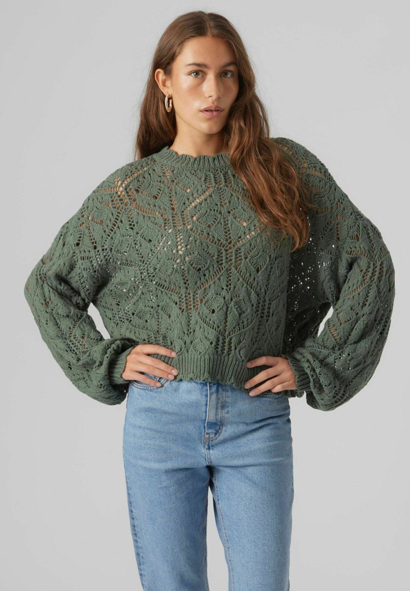 Grüner Strickpullover mit einem strukturierten Rautenmuster, weiten Ärmeln und einem gerippten Saum, kombiniert mit hellblauen, hochgeschnittenen Jeans.