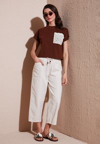 Brown cropped t-shirt with white crochet pocket, white loose-fitting pants, and brown sandals with decorative accents. Pink sunglasses worn.
