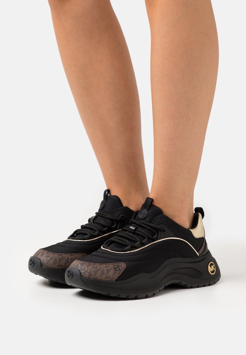 Paire de baskets épaisses noires et brunes avec des accents dorés portées aux pieds sur un fond uni.