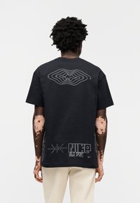 Μαύρο βαμβακερό t-shirt με λευκό αφηρημένο γραφικό στο πίσω μέρος, με λεπτομέρειες κειμένου. Κοντά μανίκια, άνετη γραμμή και μεγάλο σχεδιασμό εκτύπωσης.