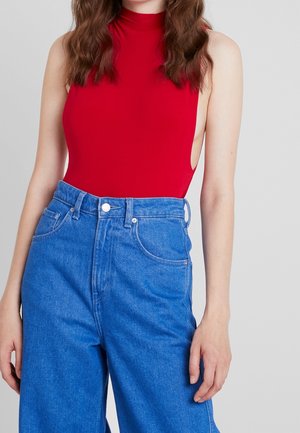 Kobieta w czerwonym topie bez rękawów z wysokim dekoltem i luźnych niebieskich dżinsach z wysokim stanem, stojąca na jasnym tle.