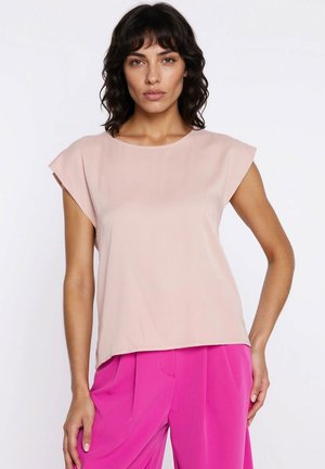 Kobieta o ciemnych kręconych włosach, ubrana w jasnoróżowy bezrękawnik i jaskrawo różowe spodnie z wysokim stanem, stojąca na neutralnym tle.