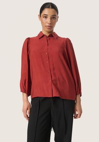 Red collared blouse with long puff sleeves, made of a smooth fabric. Features button closure and a relaxed fit, paired with black trousers.