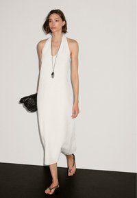 Robe maxi blanche sans manches avec un design à encolure halter. Texture lisse,coupe ample, assortie à un sac en osier noir et des tongs.