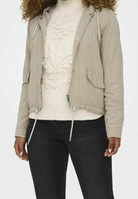 Light gray cropped jacket with a drawstring hood, zip closure, and front pockets, layered over a cable-knit white sweater. Black jeans.
