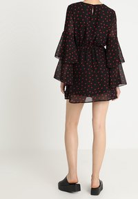 Mujer vestida con un mini vestido negro transparente con patrones de corazones rojos, mangas acampanadas en capas y zapatos de plataforma negros tipo slip-on, de pie contra un fondo blanco.