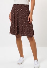 Jupe marron à motif avec une silhouette évasée, présentant un design géométrique en crème. Fabriquée à partir d'un tissu doux et texturé. Associée à des baskets blanches.