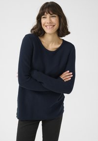 Marineblaue gerippte Langarmstrickjacke mit Rundhalsausschnitt. Weicher Stoff, lockerer Sitz und leicht verlängertes Rückenteil. Kombiniert mit schwarzen Hosen.