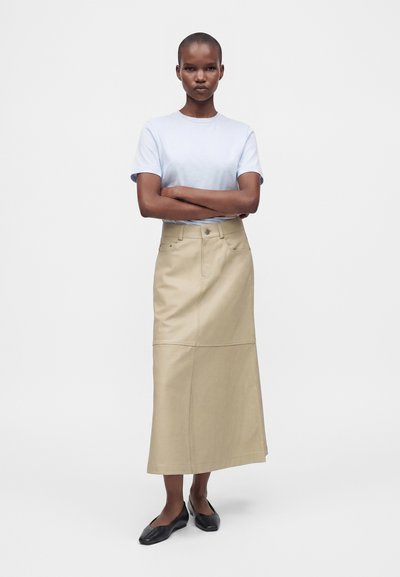 Femme aux cheveux courts portant un t-shirt bleu clair, une longue jupe beige et des chaussures plates noires, debout les bras croisés sur un fond blanc.