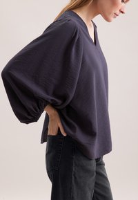 Løs, mørk lilla bluse med V-udskæring, pufærmer og struktureret stof. Sammen med højtaljede sorte jeans.