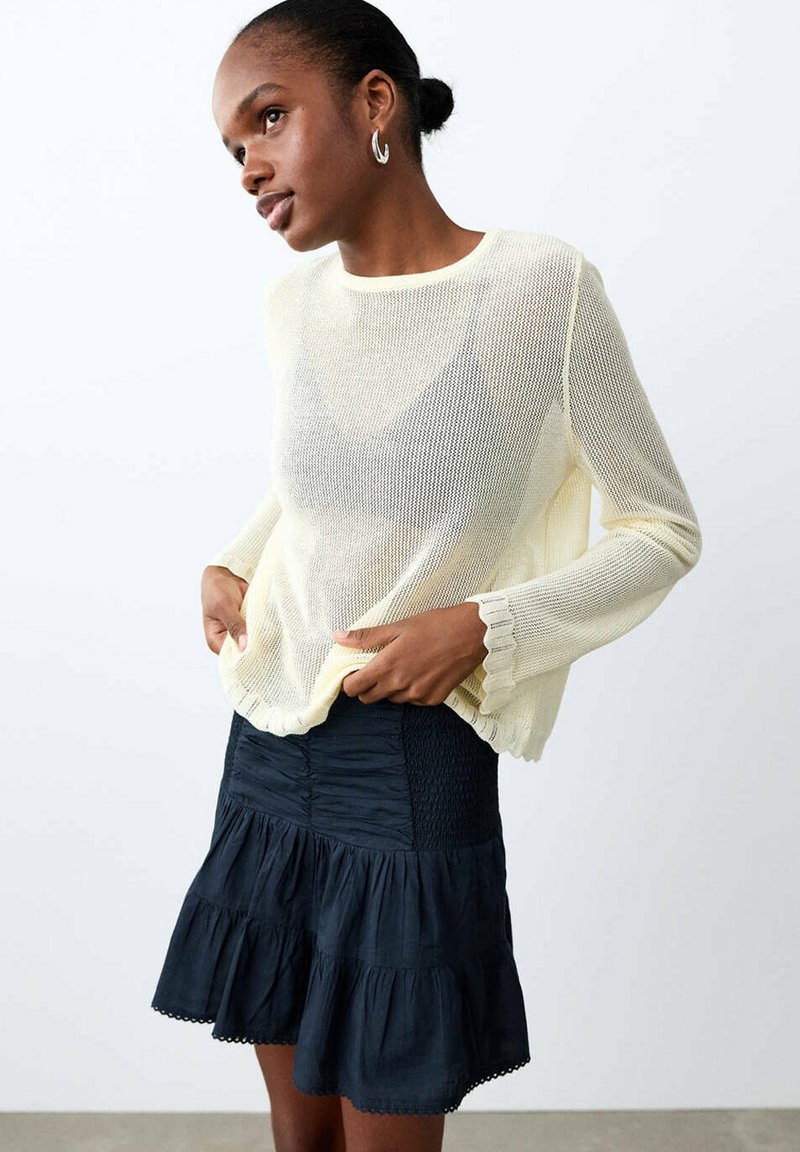 Femme portant un pull crème transparent à manches longues et une jupe noire à volants en étages, posant avec les mains près de la taille sur un fond uni.