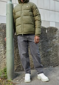 Oliwkowo-zielona pikowana kurtka na zamek, szare luźne spodnie dżinsowe oraz masywne białe sneakersy na platformie, stojąca na tle betonowej ściany.