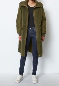 Olivgrön, lång kappa med huva och fleecefoder, dubbel fickor och tryckknappar. Bärs över en svart topp och paras med mörka jeans och vita sneakers.