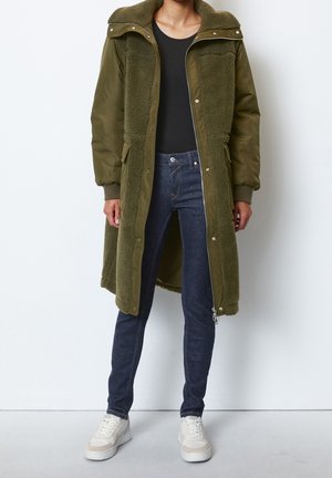 Olijfgroene, lange hoodie-jas met fleece voering, dubbele zakken en drukknoopsluitingen. Draagt over een zwart shirt en gecombineerd met donkere jeans en witte sneakers.