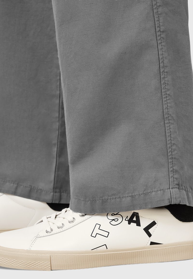 Baskets blanches avec des accents graphiques noirs, associées à un pantalon gris. Les baskets possèdent des lacets et une semelle texturée.