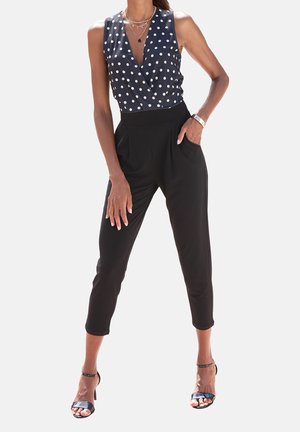 Mujer con top cruzado sin mangas azul marino con lunares, combinado con pantalones tobilleros negros de cintura alta y tacones negros con correa en el tobillo.