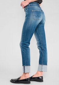 Jeans en denim bleu avec une coupe slim, présentant un ourlet roulé qui révèle un tissu intérieur contrastant. Comprend des poches arrière avec un design de point de couture distinctif.