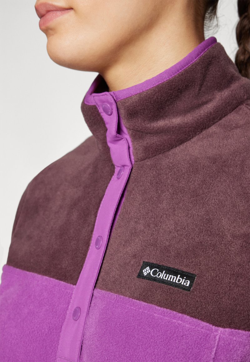 Paars en bruin fleecejack met een hoge kraag, drukknoopsluiting en een logopatch. Zachte textuur en color-blocked ontwerp.