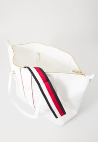 Sac à main blanc en matériau lisse, doté d'une fermeture éclair dorée, de poches intérieures et d'une bandoulière rayée rouge, blanche et bleue.