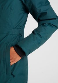 Persona con una chaqueta acolchada color azul verdoso con la mano en el bolsillo, mostrando el puño negro y el logo de la marca bordado en la manga.