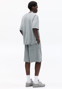 Světle šedé oversized tričko s krátkým rukávem, spárované s odpovídajícími šortkami s kapsou. Na sobě má šedé tenisky a bílé ponožky.