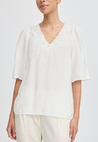 Blusa blanca con un escote en forma de V acentuado por bordados florales, mangas cortas holgadas y una tela texturizada.