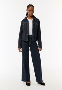 Donkerdenim jasje met bijpassende wijdlopende donkerdenim broek. Witte T-shirt eronder. Opvallende stikseldetails en knoopsluitingen.