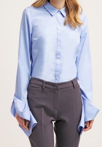 Camisa de manga larga azul claro con cuello, que presenta mangas acampanadas amplias; combinada con pantalones oscuros grises que exhiben una textura suave.