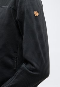 Black textured long-sleeve jacket sleeve with visible seam and small brown fox logo patch near the shoulder.