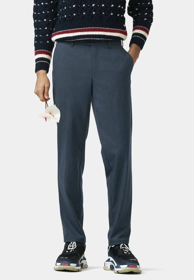 Dunkelgraue, nach Maß geschnittene Hose mit geradem Schnitt, kombiniert mit einem navyblauen Pullover mit Punkten und schwarzen Sneakers. Eine weiße Blume wird in der Hand gehalten.