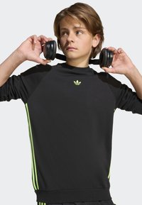 Black sweatshirt with long sleeves, ribbed cuffs, and neon green side stripes. Features a small green adidas logo on the chest. Wireless headphones held in hand.