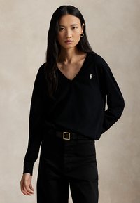 Mujer con cabello largo y negro que lleva un suéter negro de cuello en V con logo blanco y pantalones negros, de pie con una mano detrás de la espalda.