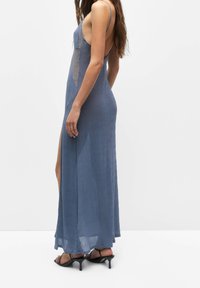 Longue robe bleue en tissu texturé avec des panneaux latéraux en dentelle et une fente haute. Caractérisée par des bretelles fines et un dos décolleté. Sandales à talons noires.