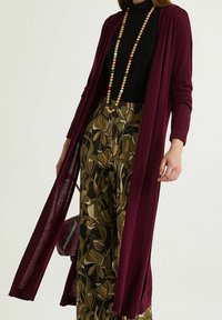 Long gilet bordeaux à devant ouvert, en tissu doux. Associé à un pantalon large à motifs verts et marrons et à un col roulé noir.