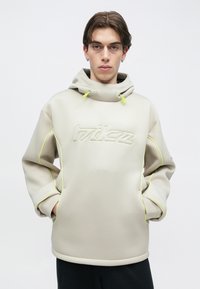 Νεαρός άνδρας που φοράει μπεζ φούτερ με φερμουάρ νέον κίτρινου χρώματος και ανάγλυφο κείμενο, στέκεται με τα χέρια στις τσέπες μπροστά σε απλό φόντο.