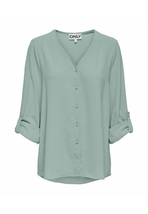 Lichtgroene blouse met lange mouwen en V-hals, knopen aan de voorkant bekleed met stof, en opgerolde mouwen die worden vastgezet met knoopbandjes.