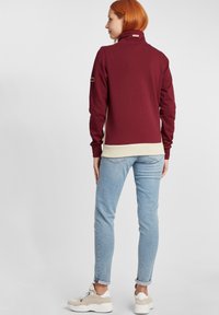 Burgunderfarget zip-up sweatshirt med høy krage, kremfarget hem og sideakenter. Kombinert med lyseblå jeans og beige sneakers.