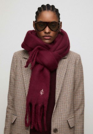 Sciarpa di lana bordeaux con estremità frangiate e logo ricamato, avvolta attorno al collo, abbinata a un blazer a pied-de-poule.
