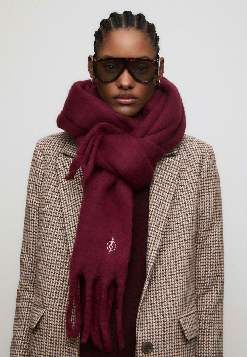 Sciarpa di lana bordeaux con estremità frangiate e logo ricamato, avvolta attorno al collo, abbinata a un blazer a pied-de-poule.