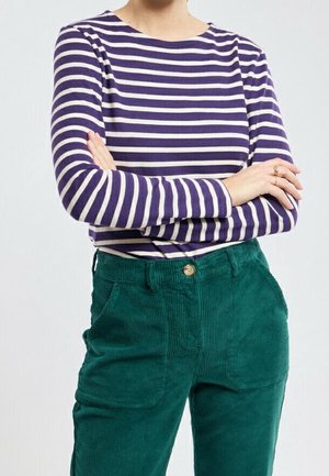 Person with arms crossed wearing a purple and white striped long-sleeve shirt and green corduroy pants with a button closure.