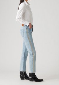 Světle modré džíny s rovnými nohavicemi, zdobené stříbrnými postranními pruhy, ve spojení s černými kotníkovými botami a bílou košilí na knoflíky.