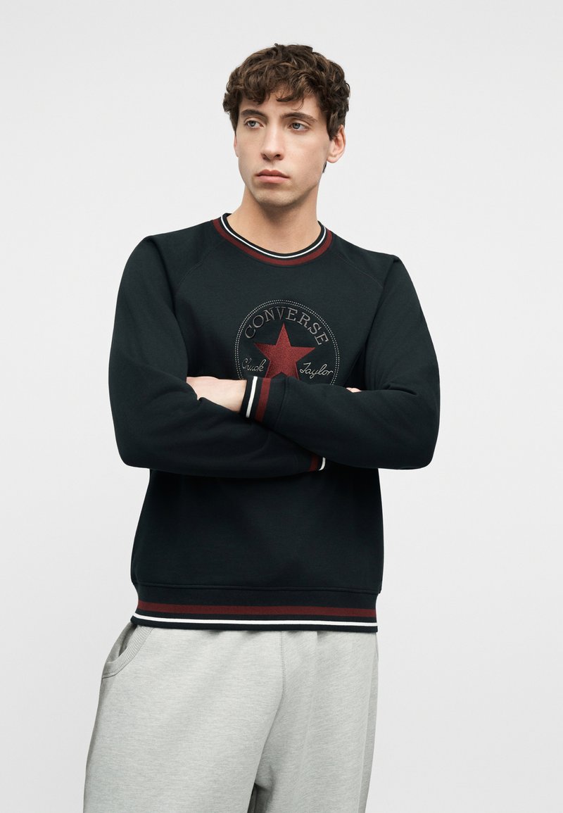 Young man with crossed arms wearing black Converse sweatshirt with red star logo and light gray sweatpants, standing against plain background.