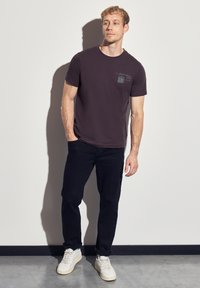 Tmavě fialové tričko s krátkým rukávem a grafikou na hrudi, spárované s černými kalhotami a bílými vysokými teniskami. Hladká bavlněná textura.