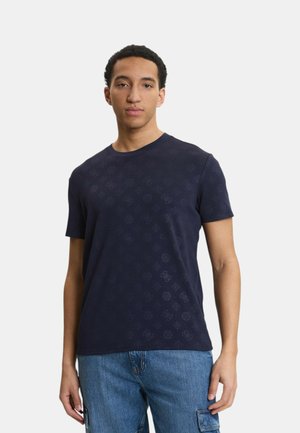 Giovane uomo che indossa una t-shirt blu navy con motivo e jeans blu, in piedi davanti a uno sfondo semplice.