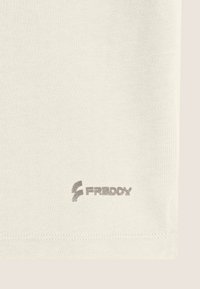 Camiseta de color beige claro con un tejido liso y texturizado. Presenta un logo plateado con el texto "FREDDY" cerca del dobladillo inferior.