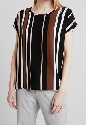 Femme portant un chemisier à manches courtes à rayures verticales noires, marron, blanches et roses, associé à un pantalon gris clair sur un fond uni.