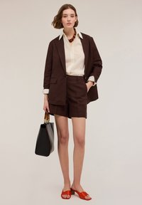 Brun skräddarsydd kavaj och shorts i slät tyg, kombinerat med en ljus knapp-up skjorta. Tillbehör inkluderar ett tjockt halsband och en svart väska.